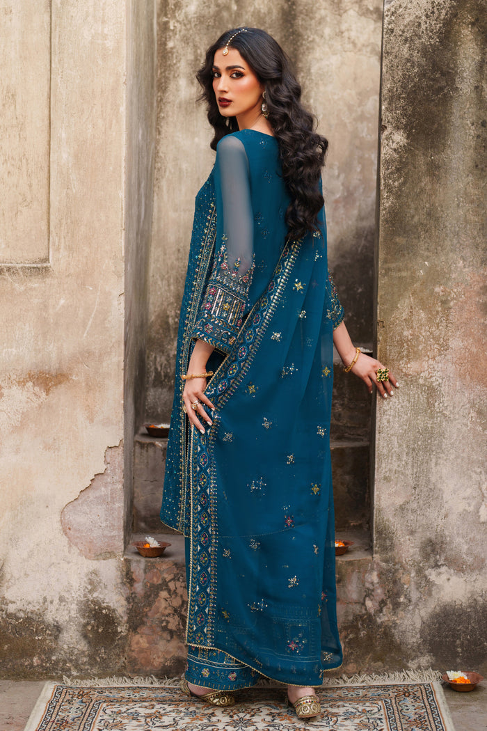 Woman in a blue saree with intricate patterns standing against a textured wall.