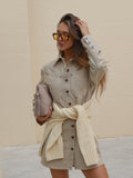 Woman wearing a beige denim dress and sweater with sunglasses, holding a clutch against a beige wall.