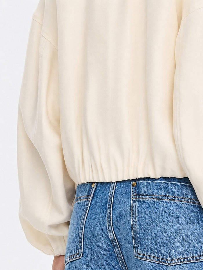 Close-up of a person wearing a cream-colored long-sleeve top with a gathered elastic hem, paired with blue denim jeans.
