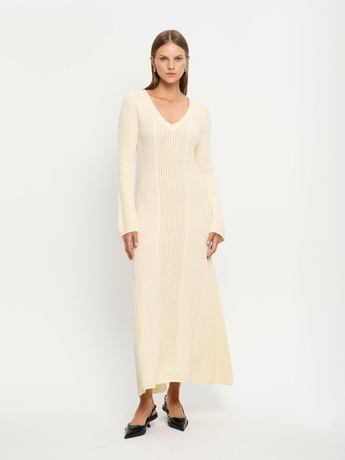 A female model wearing a beige Elliot Knit Dress with a V-neckline and cable knit stitch detailing, complete with a waist tie.
