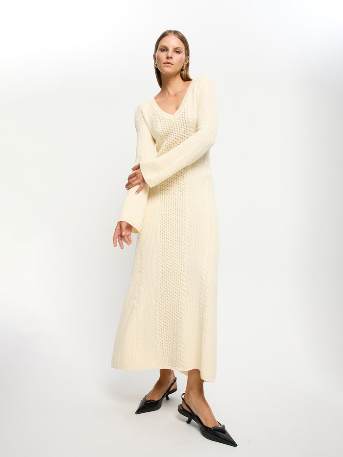 A female model wearing a beige Elliot Knit Dress with a V-neckline and cable knit stitch detailing, paired with black shoes.