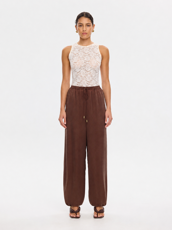 A model stands against a plain white studio background wearing a sleeveless white lace top and wide-leg brown pants with a drawstring waist.