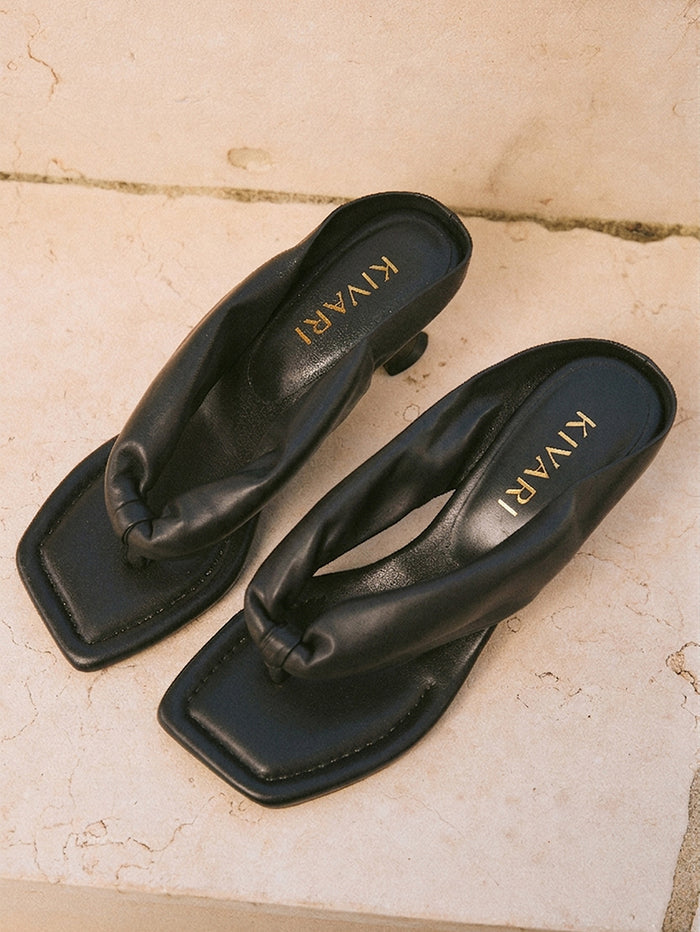 Pair of black heels with 'KIVARI' branding on a beige stone surface
