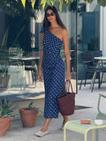 Woman in a blue polka dot dress standing outdoors with potted plants and tables in the background.