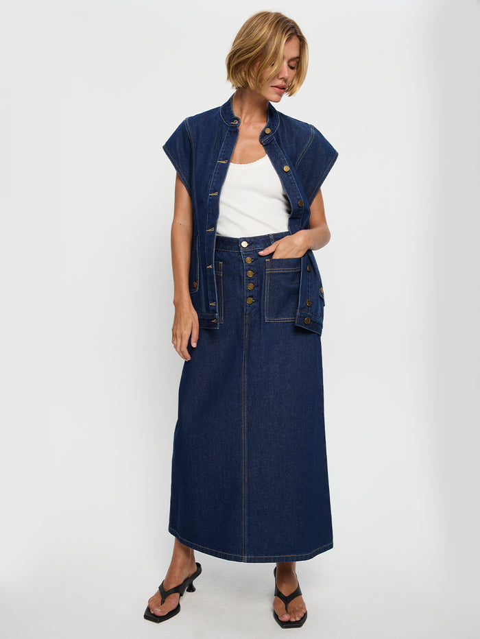 A woman standing in a studio setting, wearing a navy blue denim midi skirt with contrast topstitch detailing and patch pockets, along with a white top and black sandals.