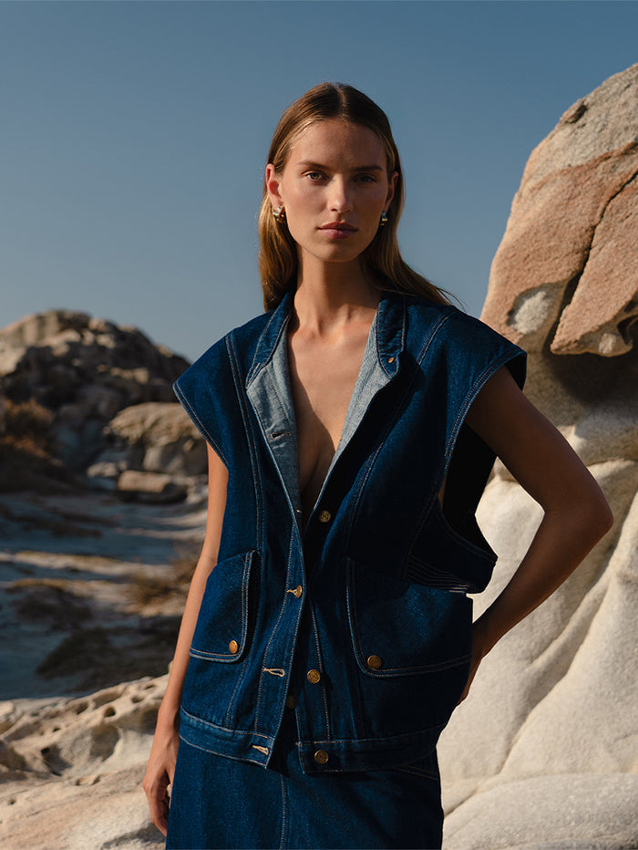 A person wearing a blue denim vest with front patch pockets and contrast top stitching, standing near a rocky coast.