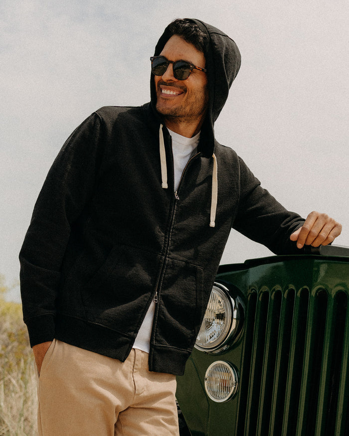 fit model leaning against a car wearing The Apres Zip Hoodie in Black Indigo Terry