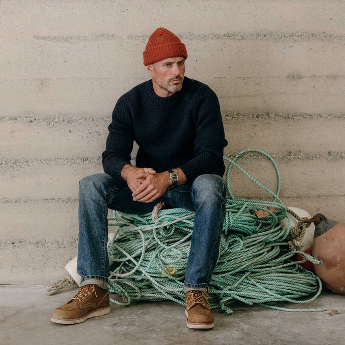 fit model sitting on a bunch of rope wearing The Fisherman Sweater in Dark Navy Merino