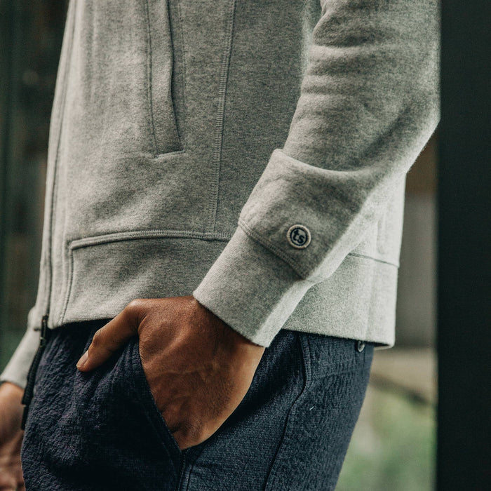 our fit model wearing The Heavy Bag Bomber in Heather Grey Fleece closeup with right hand in pocket sshowcasing embroidered TS logo on cuff