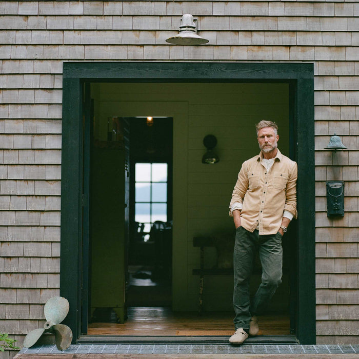 fit model leaning against a house wearing The Utility Shirt in Camel Corduroy